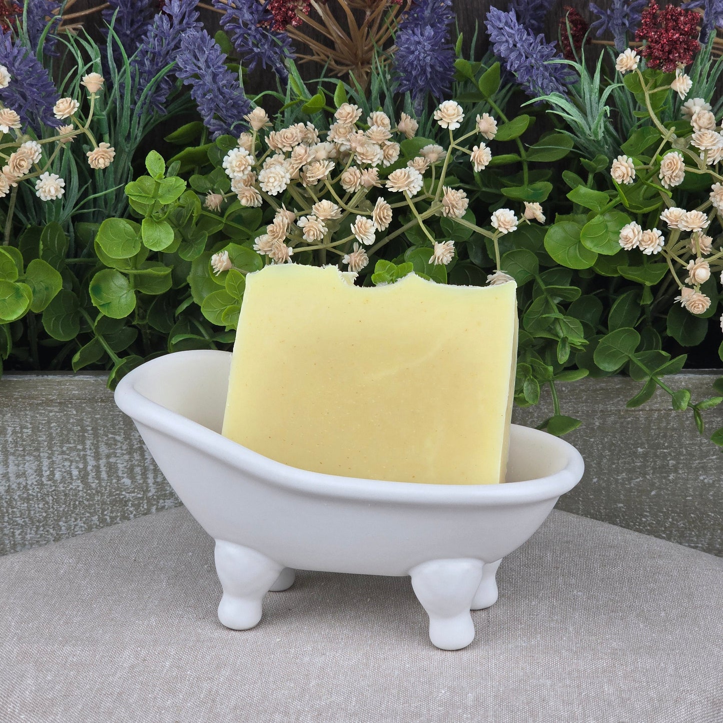 The Lavender + Peppermint Handmade Natural Artisan Soap by Jarhead Aromatics rests in a small white ceramic bathtub dish, with faux purple, white, and red flowers and green leaves in the background.