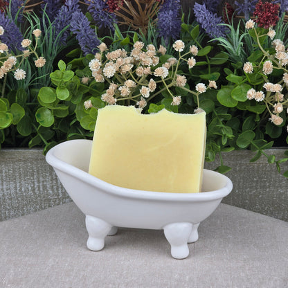 The Lavender + Peppermint Handmade Natural Artisan Soap by Jarhead Aromatics rests in a small white ceramic bathtub dish, with faux purple, white, and red flowers and green leaves in the background.