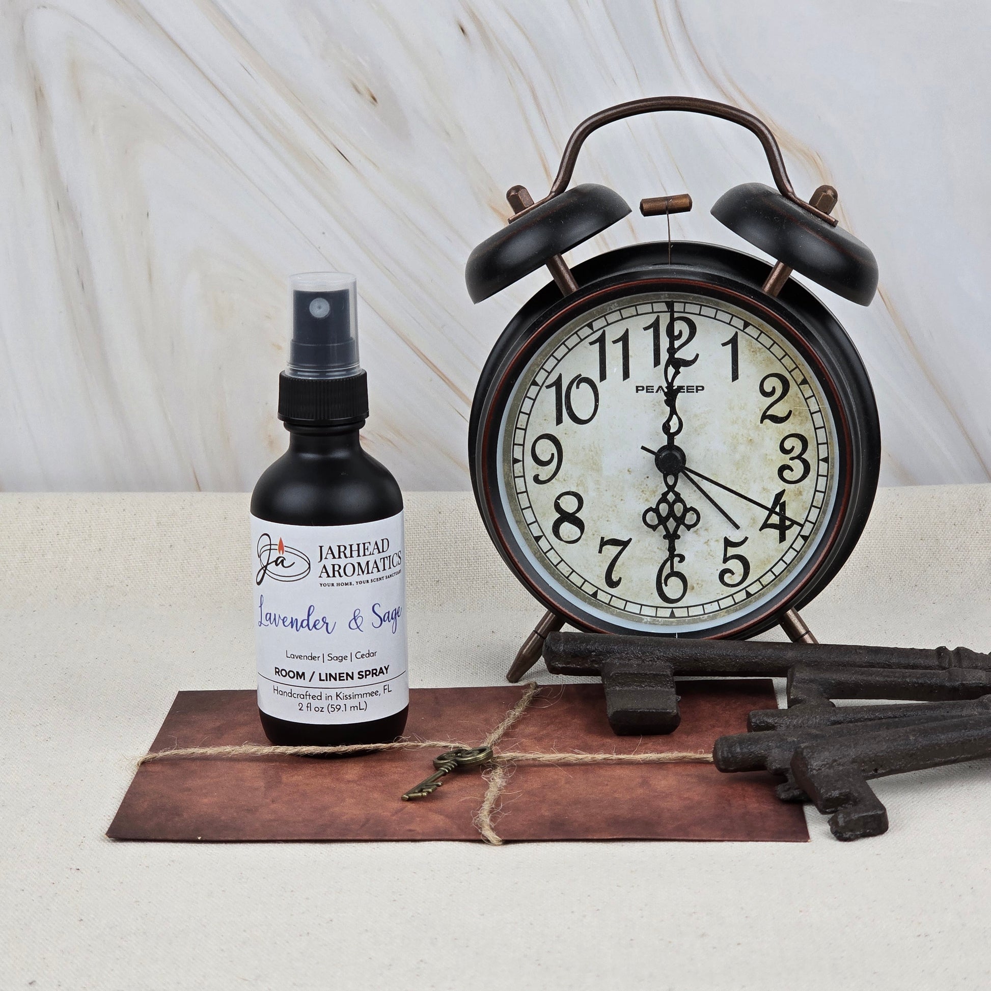 A bottle of Jarhead Aromatics Lavender & Sage Room & Linen Spray | Herbal Spa Home Fragrance Spray rests beside a vintage-style alarm clock, three large metal keys, a small ornate key, and a string-tied brown envelope on a light surface.