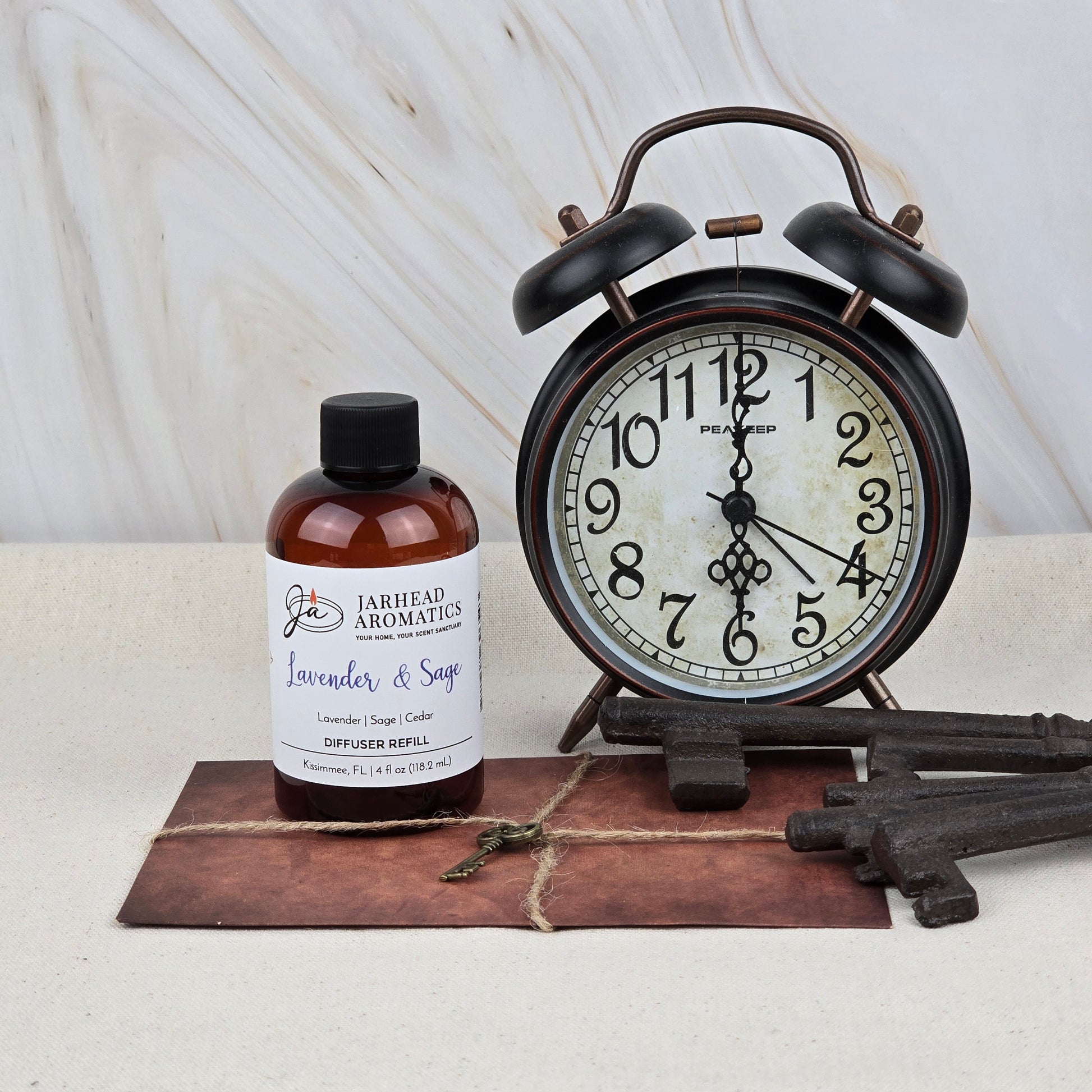 A bottle of Jarhead Aromatics Lavender & Sage Diffuser Refill | Lavender, Sage & Sandalwood Home Fragrance Oil Refill is placed by a vintage black alarm clock, large keys, and a brown envelope on a light stone surface with a white marbled background.