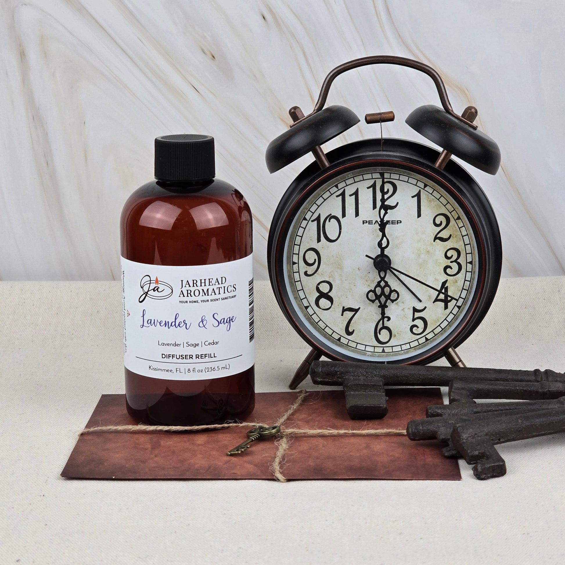 A bottle of Jarhead Aromatics Lavender & Sage Diffuser Refill | Lavender, Sage & Sandalwood Home Fragrance Oil Refill rests by a vintage black alarm clock, old keys, and a postcard, creating a tranquil spa-inspired vibe with its calming scent.