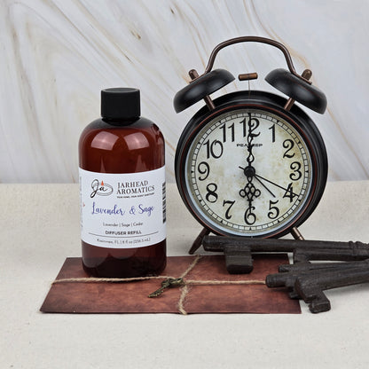 A bottle of Jarhead Aromatics Lavender & Sage Diffuser Refill | Lavender, Sage & Sandalwood Home Fragrance Oil Refill rests by a vintage black alarm clock, old keys, and a postcard, creating a tranquil spa-inspired vibe with its calming scent.
