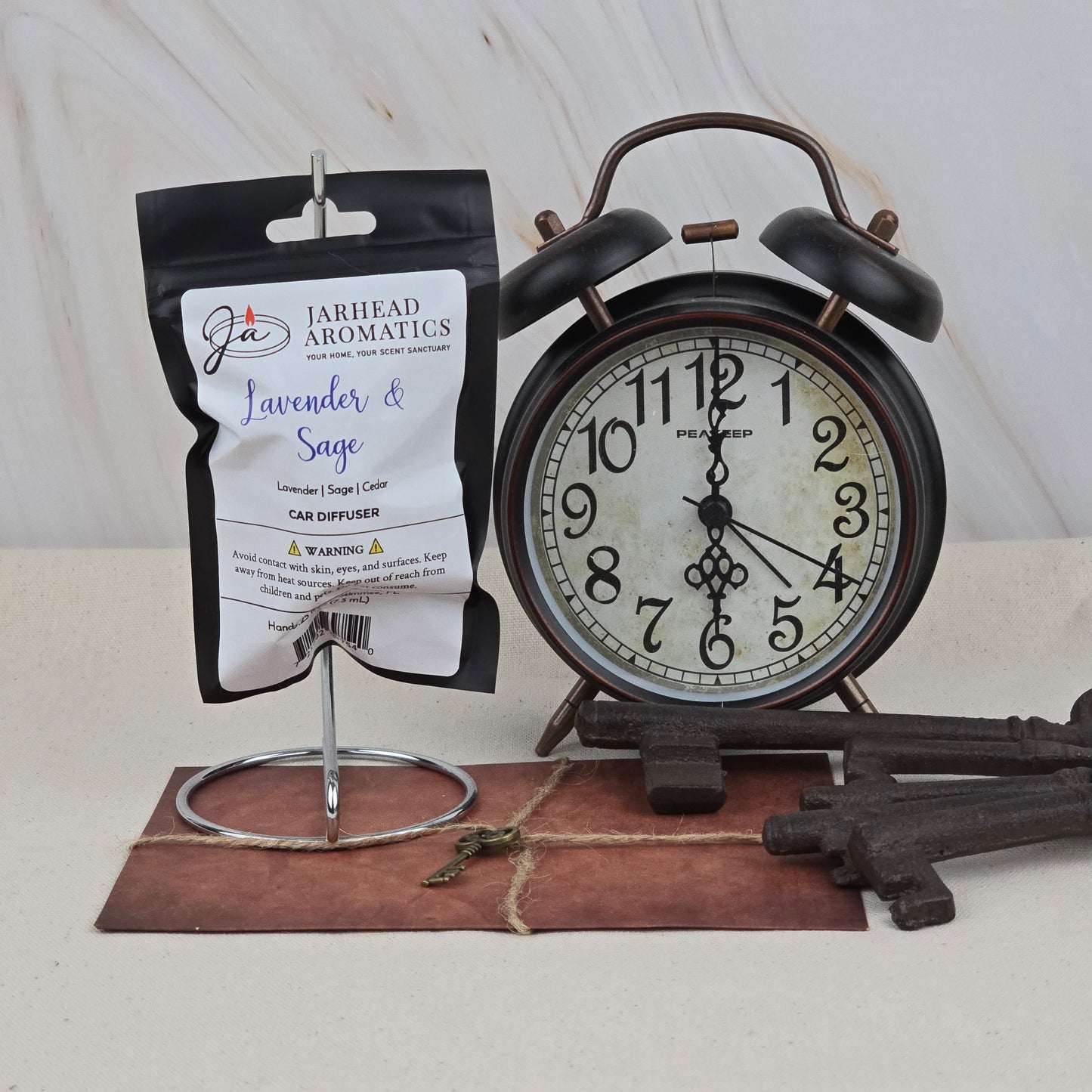 A black alarm clock sits beside a Jarhead Aromatics Lavender & Sage Car Diffuser | Herbal Spa & Sandalwood Car Air Freshener, which hangs on a metal stand with vintage keys and a brown envelope on a light surface.