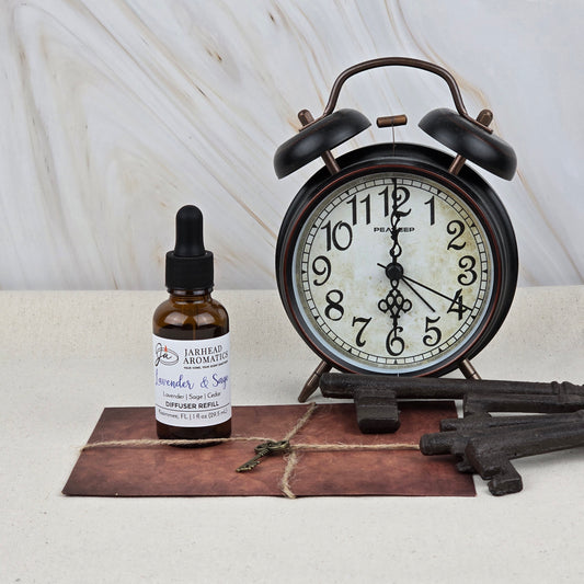 A vintage black alarm clock, antique keys, and a Jarhead Aromatics Lavender & Sage Car Diffuser Refill create a calming vibe on a light surface with marble background, evoking an herbal spa and sandalwood car fragrance ambiance.
