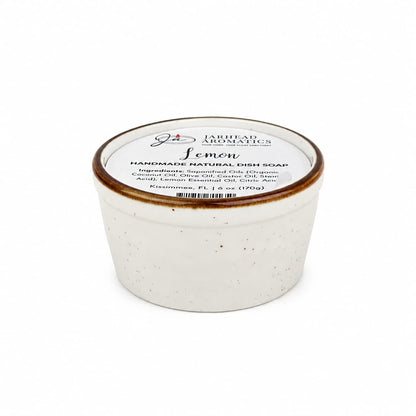 A white ceramic container with a lid labeled Lemon Handmade Natural Solid Dish Soap from Jarhead Aromatics, featuring a plant-based formula with lemon essential oil and displaying ingredients and brand information on the top.