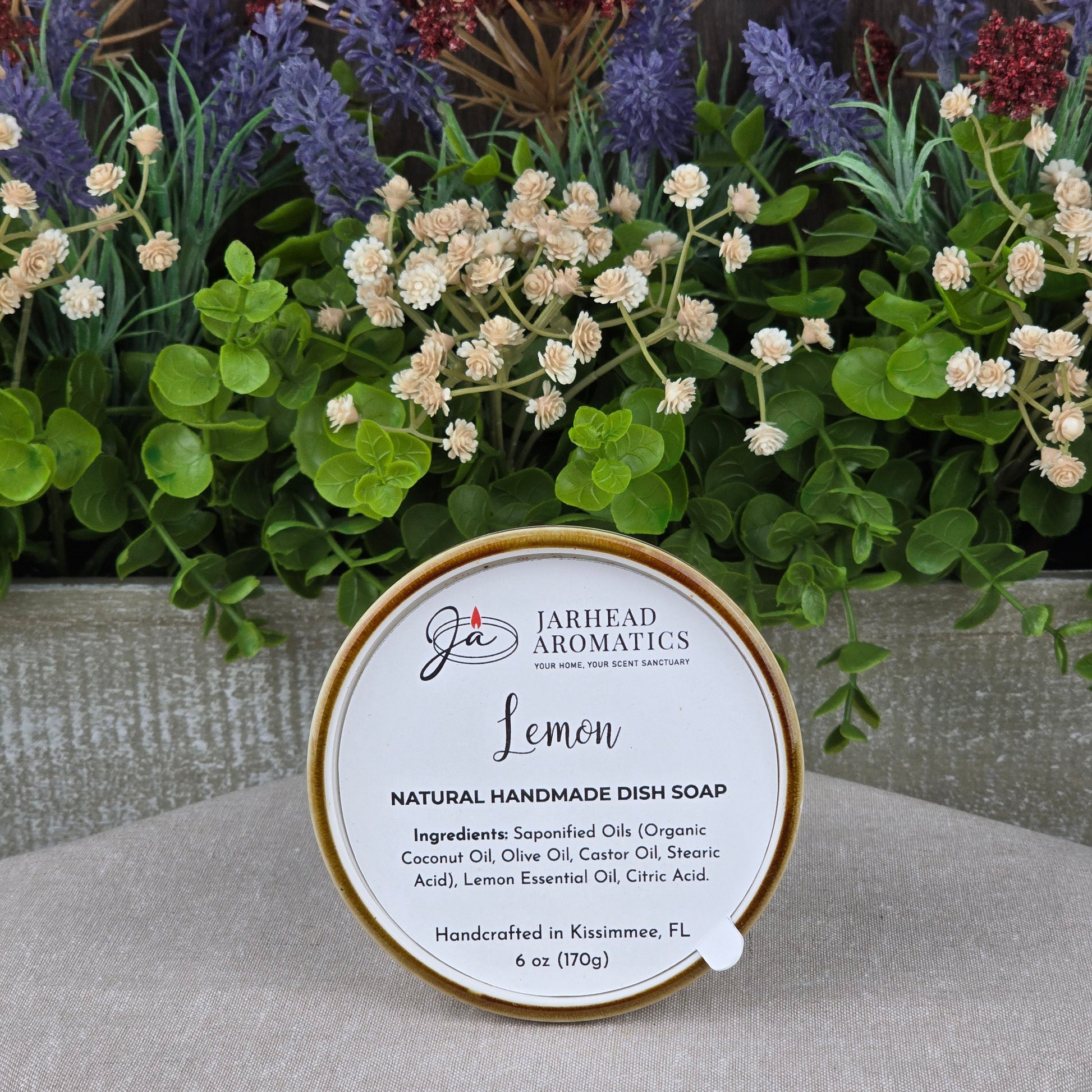 A round tin of Jarhead Aromatics Lemon Handmade Natural Solid Dish Soap sits before lush artificial flowers, its label listing ingredients and noting it is handcrafted in Kissimmee, FL.
