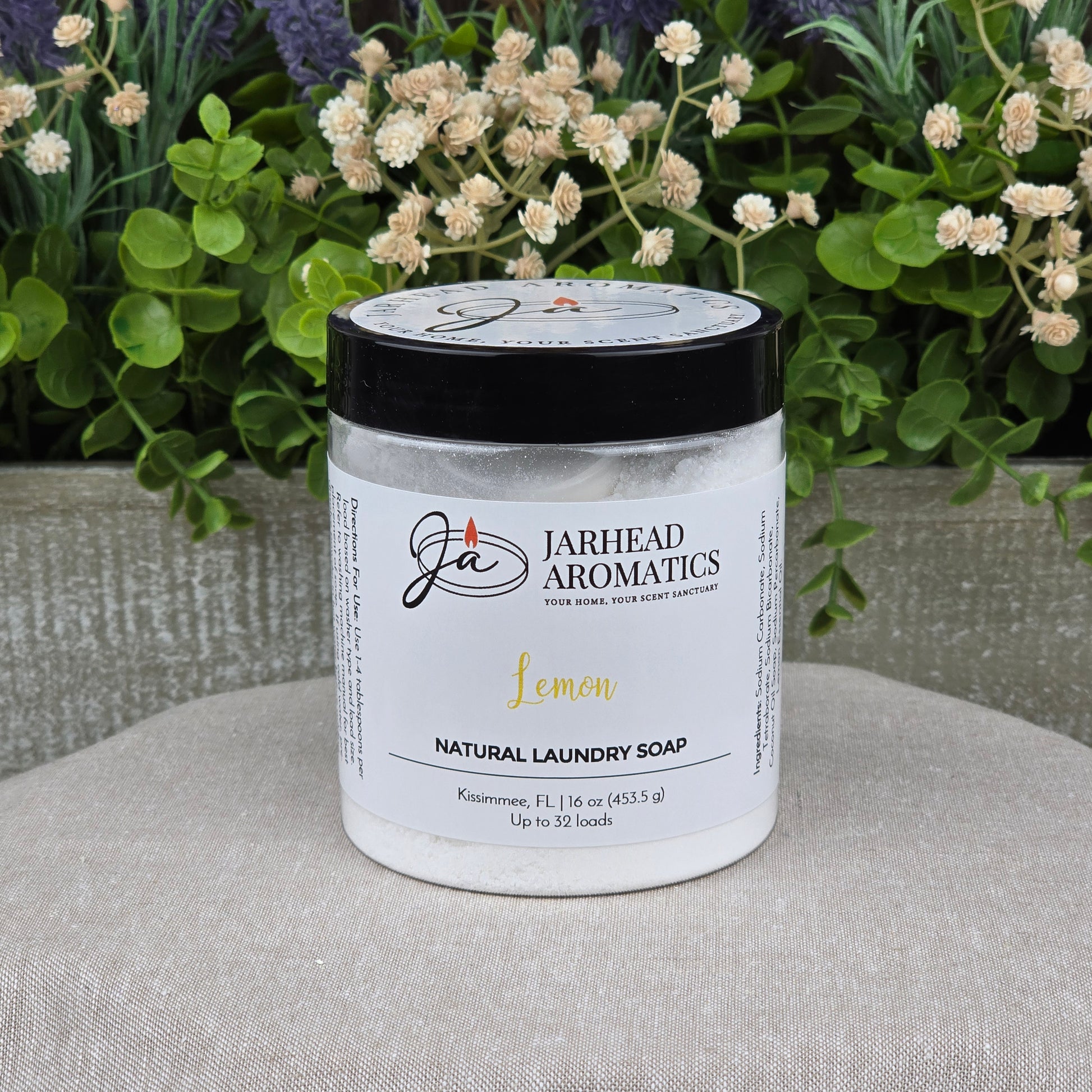 A jar of Jarhead Aromatics Lemon Handmade Natural Laundry Soap sits on a cushion, framed by greenery and white flowers. The non-toxic citrus detergent powder is 16 oz and lasts for up to 32 loads.