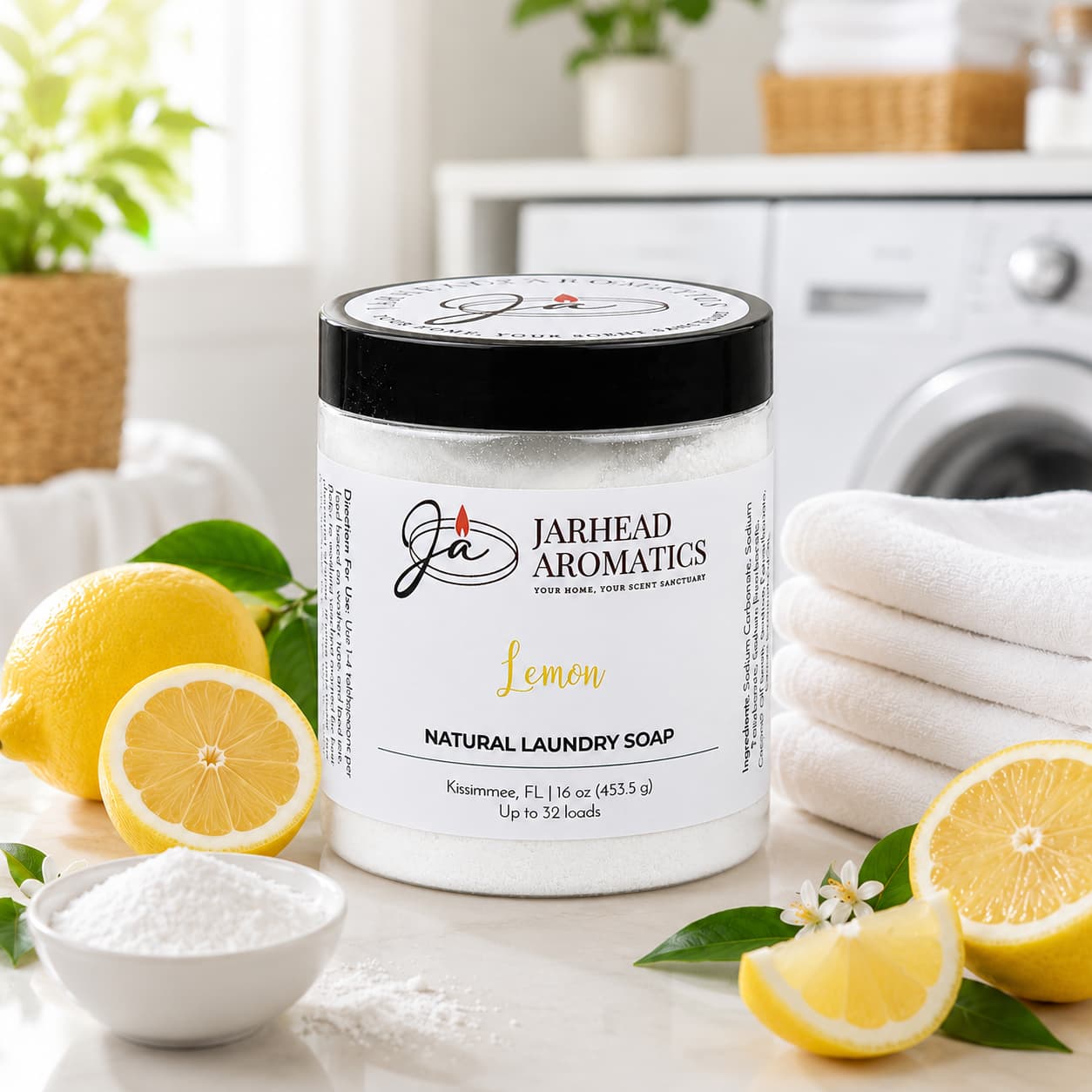 A container of Jarhead Aromatics Lemon Handmade Natural Laundry Soap | Powdered Detergent with Lemon Essential Oil rests on a countertop, accompanied by vibrant fresh lemons, a bowl filled with artisanal powdered detergent, and stacks of neatly folded towels. Behind it, a washing machine and a flourishing green plant add to the bright and invigorating atmosphere.