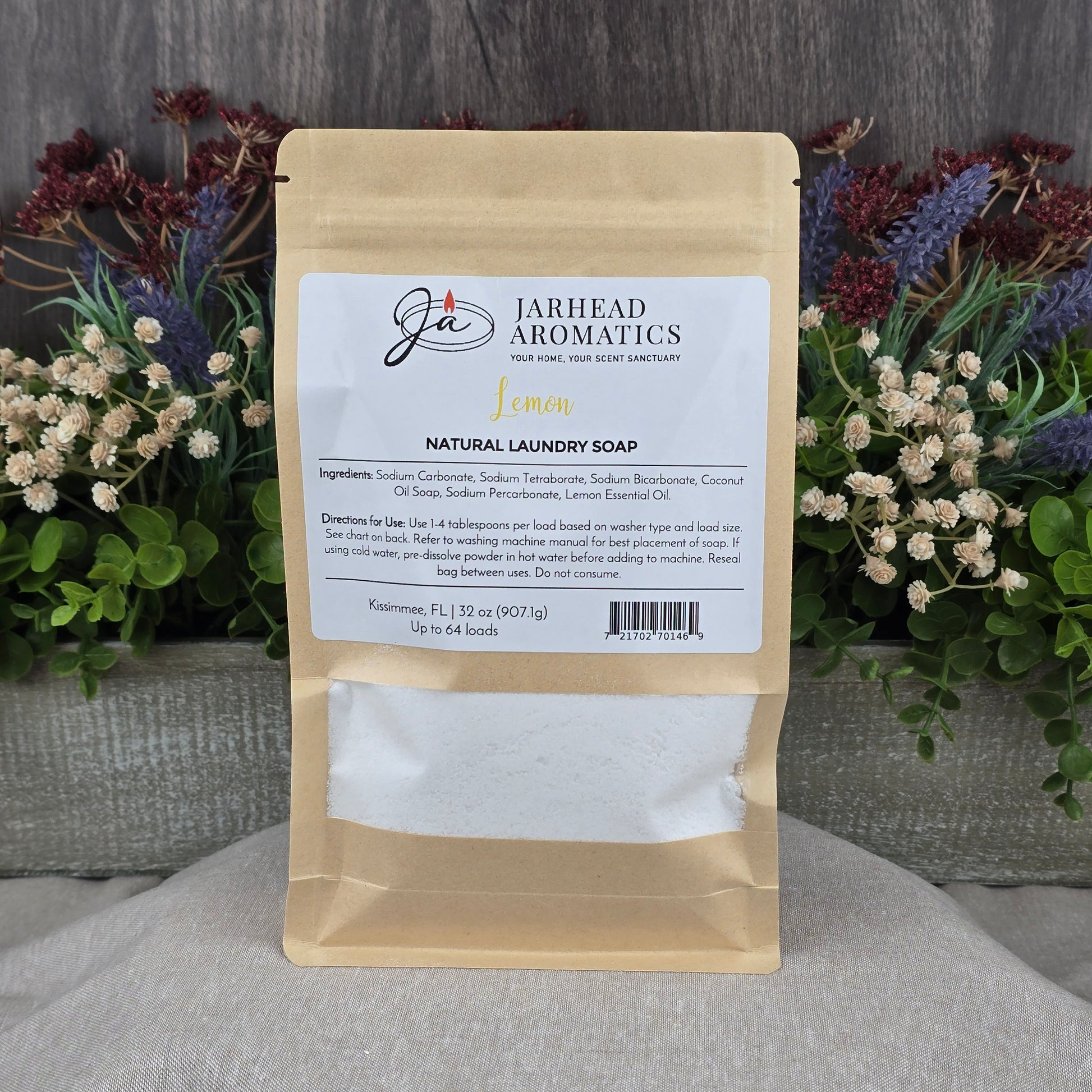 A brown paper pouch of Jarhead Aromatics Lemon Handmade Natural Laundry Soap | Non-Toxic Citrus Laundry Detergent Powder rests on a gray surface, with a colorful floral arrangement in the background. The label shows ingredients and directions.