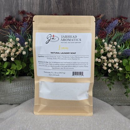 A brown paper pouch of Jarhead Aromatics Lemon Handmade Natural Laundry Soap | Non-Toxic Citrus Laundry Detergent Powder rests on a gray surface, with a colorful floral arrangement in the background. The label shows ingredients and directions.