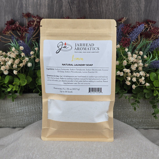 A brown paper pouch of Jarhead Aromatics Lemon Handmade Natural Laundry Soap | Non-Toxic Citrus Laundry Detergent Powder rests on a gray surface, with a colorful floral arrangement in the background. The label shows ingredients and directions.
