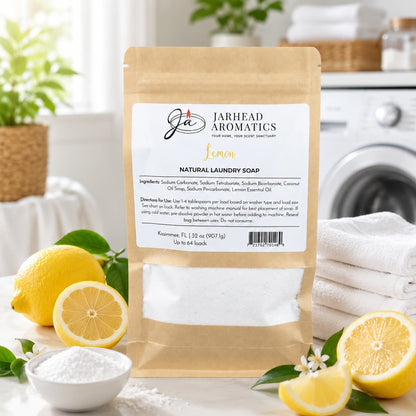 A brown paper bag labeled "Lemon Handmade Natural Laundry Soap | Powdered Detergent with Lemon Essential Oil" from Jarhead Aromatics sits on a table alongside fresh lemons, a towel, and a scoop of powder in a bright, airy room.