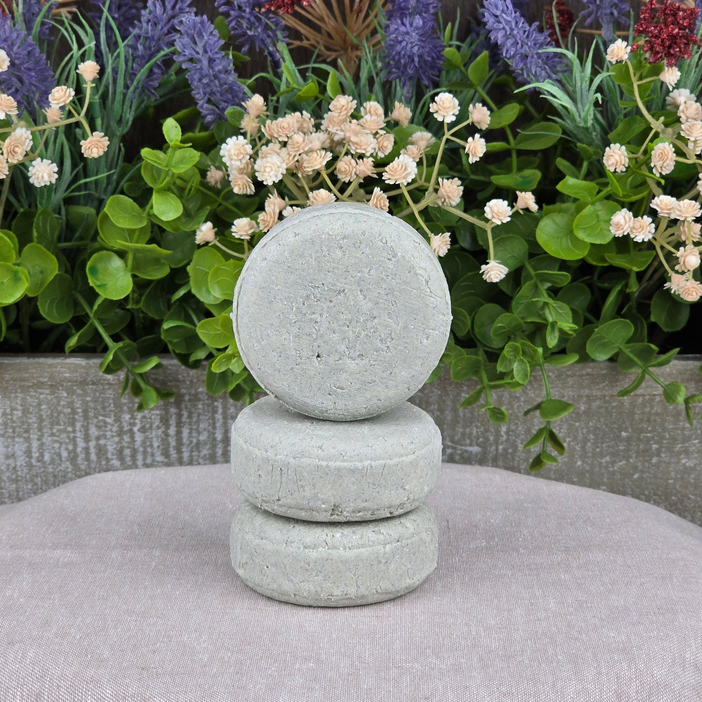 Three Lemongrass Handmade Shampoo & Conditioner Bars by Jarhead Aromatics are stacked on a light fabric, with greenery and flowers in the background—a perfect all-natural, zero-waste haircare choice.