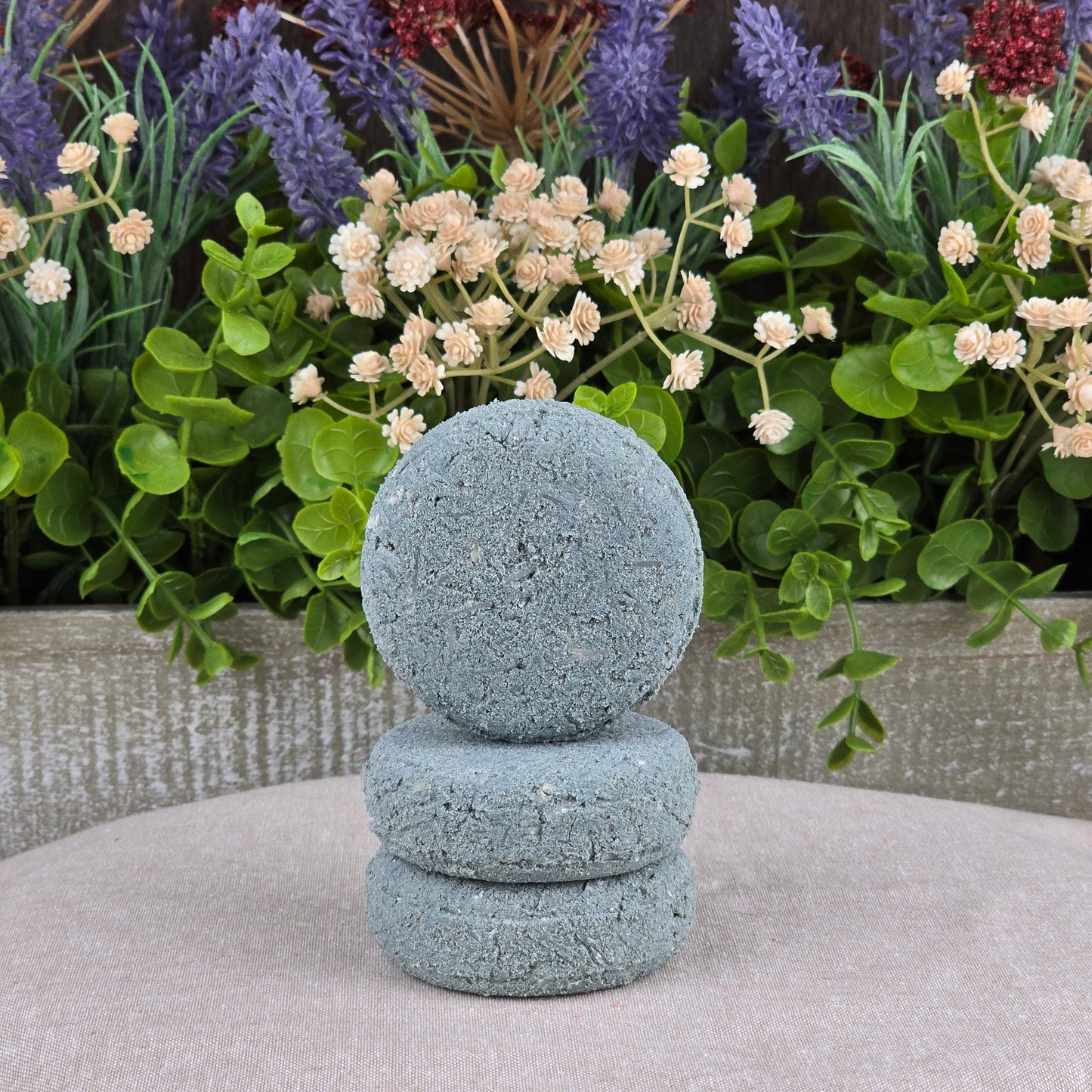 Three round, blue-gray Lemongrass Handmade Shampoo & Conditioner Bars by Jarhead Aromatics are stacked on a light, textured surface in front of green leaves and purple and white flowers—perfect for zero-waste hair care.