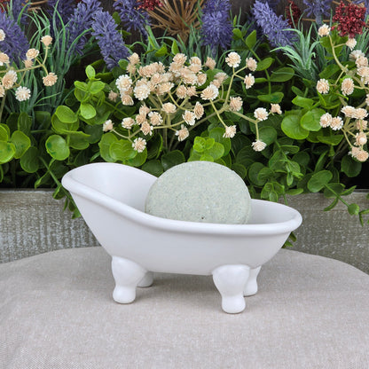 A small white ceramic bathtub dish holds the Lemongrass Handmade Shampoo & Conditioner Bar by Jarhead Aromatics. The round, pale green bar sits on a beige surface, with green plants and white and purple flowers in the background.