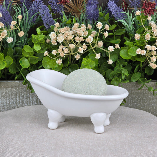 A small white ceramic bathtub dish holds the Lemongrass Handmade Shampoo & Conditioner Bar by Jarhead Aromatics. The round, pale green bar sits on a beige surface, with green plants and white and purple flowers in the background.