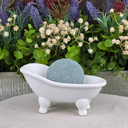 The Lemongrass Handmade Shampoo & Conditioner Bar from Jarhead Aromatics, round and blue, is displayed in a small white clawfoot bathtub dish with green and purple artificial flowers in the background.