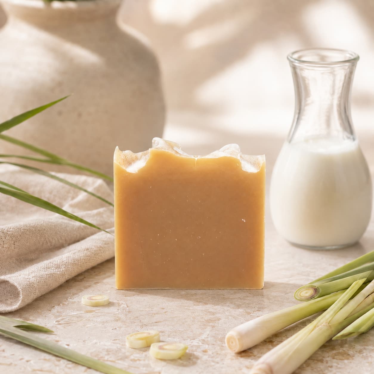 A bar of Jarhead Aromatics Lemongrass Handmade Natural Goat Milk Artisan Soap stands upright on a stone surface, accompanied by fresh lemongrass stalks, a small glass carafe of milk, and a ceramic vase in the background. Bright citrus-herbal notes are suggested as soft natural light highlights the scene.