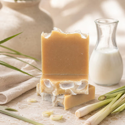 Two bars of Jarhead Aromatics Lemongrass Handmade Natural Goat Milk Artisan Soap are stacked on a marble surface, surrounded by fresh lemongrass, a beige towel, and a glass jar of milk. Soft natural light highlights the artisanal quality and bright citrus-herbal fragrance of these soothing goat milk soaps.