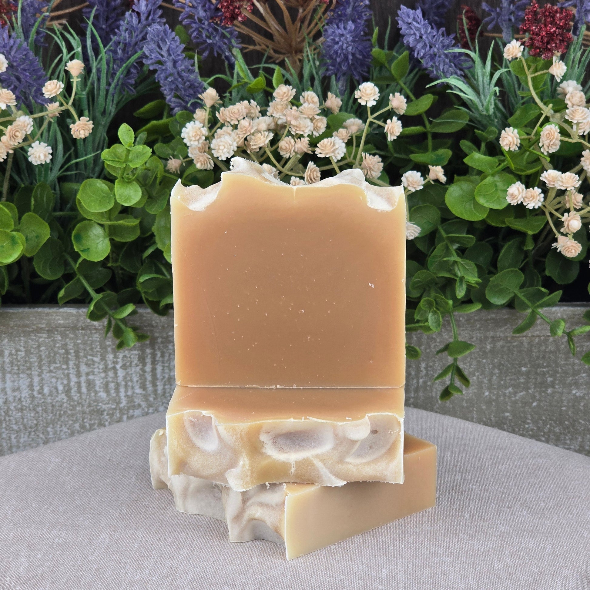Two Jarhead Aromatics soap bars, including the Lemongrass Handmade Natural Goat Milk Artisan Soap with a wavy edge, rest on light fabric. Lush greenery and dried purple, red, and white flowers create a vibrant backdrop.