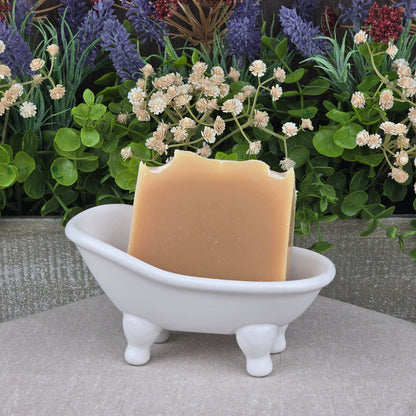 A rectangular bar of Jarhead Aromatics Lemongrass Handmade Natural Goat Milk Artisan Soap with a bite mark rests in a small white clawfoot bathtub soap dish, with artificial green, purple, and red plants and flowers behind it.
