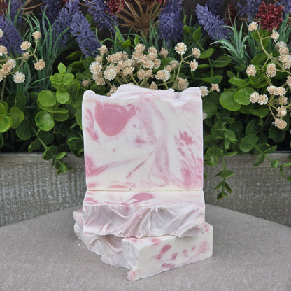 Two bars of Jarhead Aromatics Magnolia + Lavender Handmade Coconut Milk Artisan Soap, with aloe and shea butter, are stacked before vibrant artificial flowers and lush leaves on a gray textured surface, adding elegance to the setting.