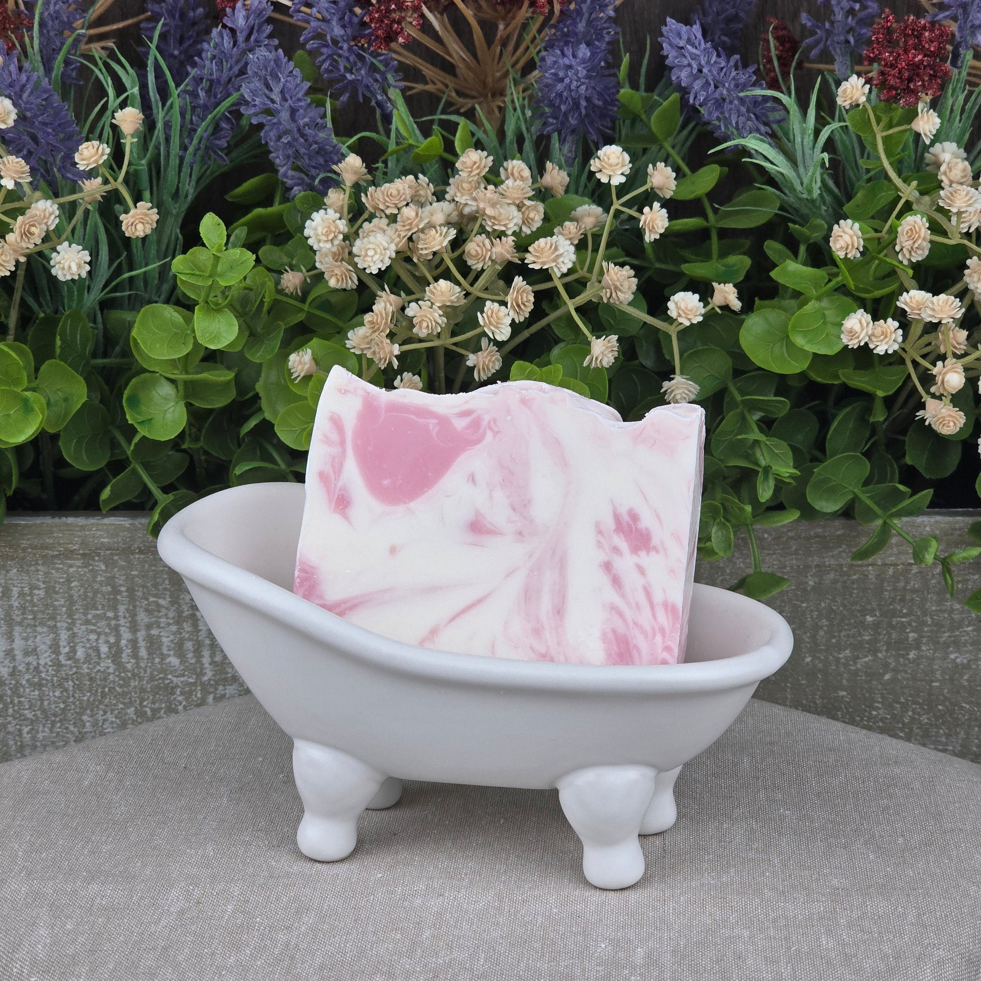 A Magnolia + Lavender Handmade Coconut Milk Artisan Soap by Jarhead Aromatics rests in a white ceramic bathtub, set against artificial green plants and colorful flowers.