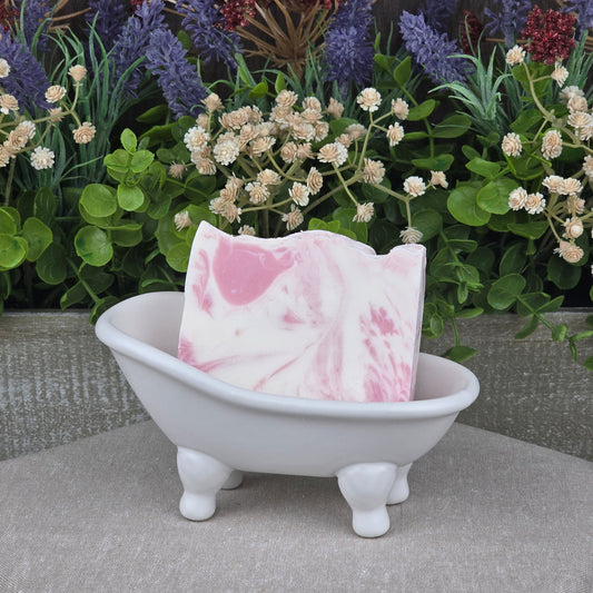 A Magnolia + Lavender Handmade Coconut Milk Artisan Soap by Jarhead Aromatics rests in a white ceramic bathtub, set against artificial green plants and colorful flowers.