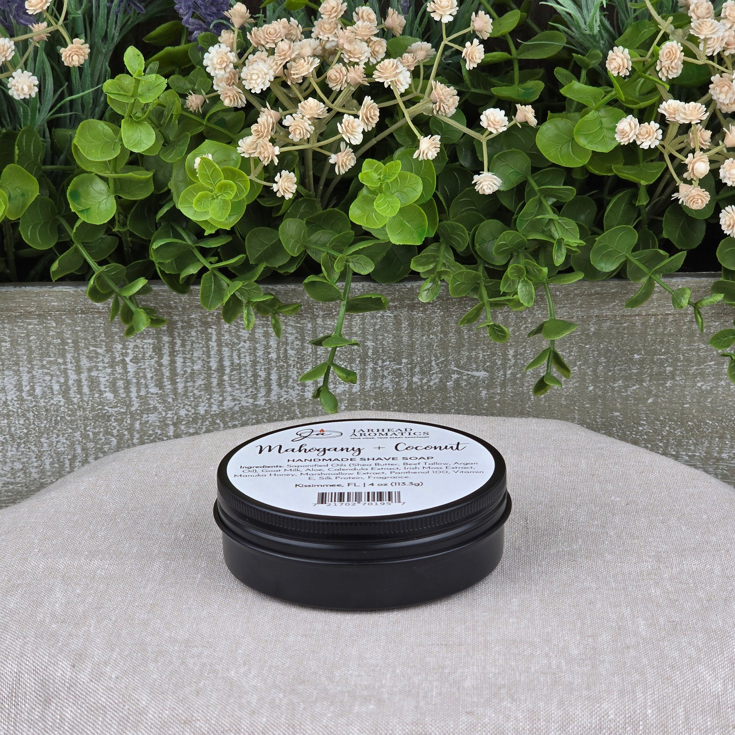 A black round container of Jarhead Aromatics Mahogany + Coconut Handmade Shave Soap | Beef Tallow & Goat Milk sits on a beige surface, with green plants and white flowers in the background, showcasing this artisan shaving soap.