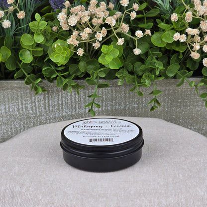 A black round container of Jarhead Aromatics Mahogany + Coconut Handmade Shave Soap | Beef Tallow & Goat Milk sits on a beige surface, with green plants and white flowers in the background, showcasing this artisan shaving soap.