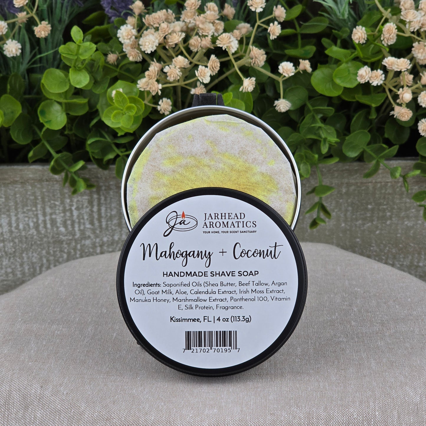 A round container of Jarhead Aromatics Mahogany + Coconut Handmade Shave Soap | Beef Tallow & Goat Milk is displayed on a beige surface, with blurred green plants and white flowers in the background. The label lists ingredients and product details.