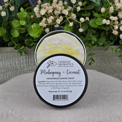 A round container of Jarhead Aromatics Mahogany + Coconut Handmade Shave Soap | Beef Tallow & Goat Milk is displayed on a beige surface, with blurred green plants and white flowers in the background. The label lists ingredients and product details.