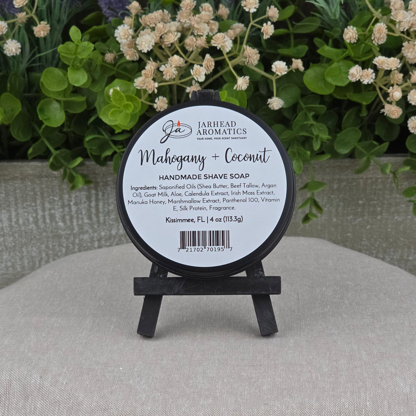 A round tin of Jarhead Aromatics Mahogany + Coconut Handmade Shave Soap with beef tallow and goat milk rests on a black easel, accented by greenery and flowers. The label showcases its natural ingredients and product details.