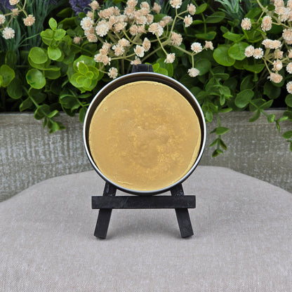 A round tin of Jarhead Aromatics Mahogany + Coconut Handmade Shave Soap rests on a black easel atop gray fabric, with green leaves and white flowers in the background.