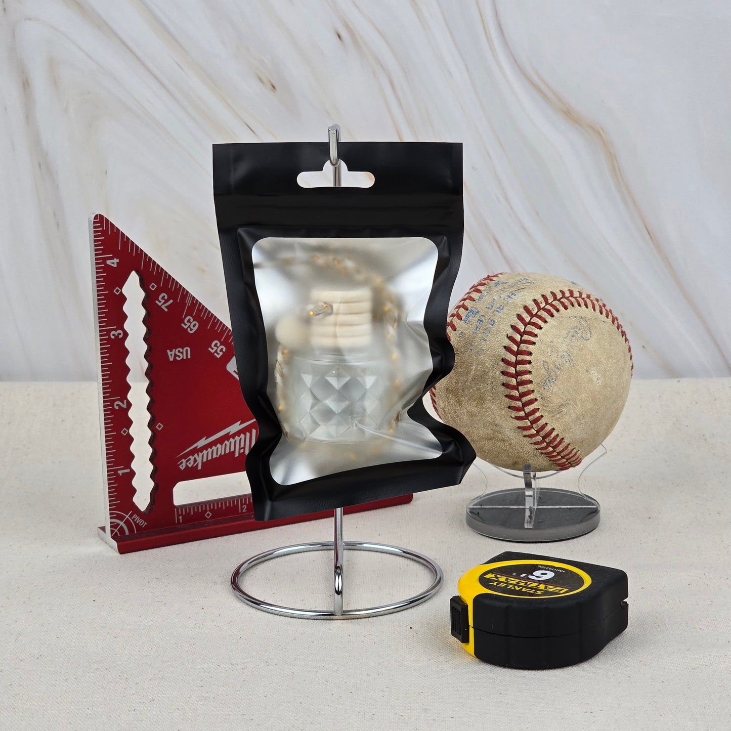 A resealable black pouch, red triangular ruler, worn baseball on a stand, and yellow tape measure are arranged on a marble-patterned background, highlighting Jarhead Aromatics' The Lumberjack Car Diffuser—an outdoorsy cedar, oak & mahogany air freshener.