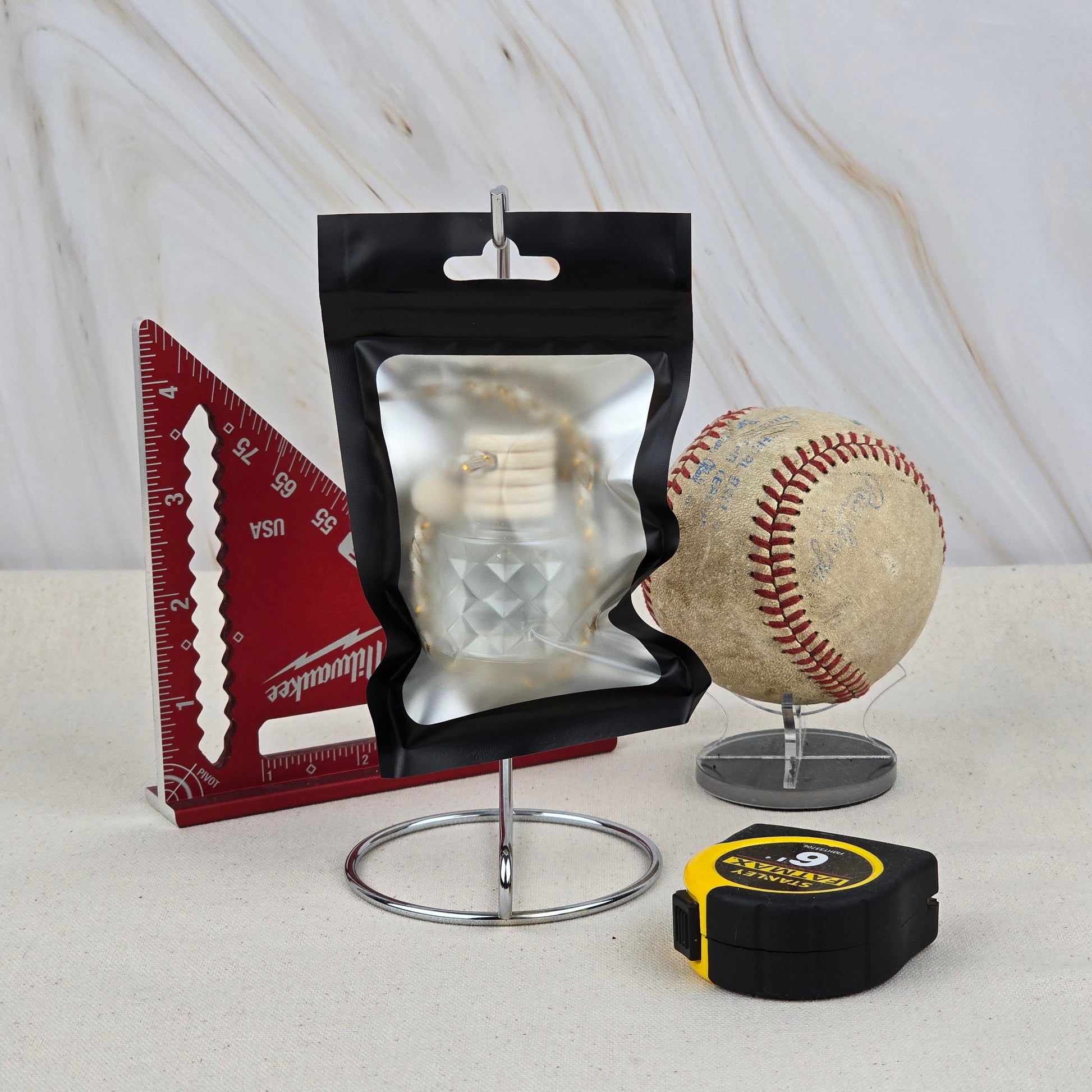 A resealable black pouch, red triangular ruler, worn baseball on a stand, and yellow tape measure are arranged on a marble-patterned background, highlighting Jarhead Aromatics' The Lumberjack Car Diffuser—an outdoorsy cedar, oak & mahogany air freshener.