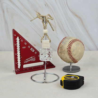 A red triangular ruler, a baseball on a stand, a black and yellow tape measure, and Jarhead Aromatics’ The Lumberjack Car Diffuser (Cedar, Oak & Mahogany) are arranged on a light surface against a marbled background.