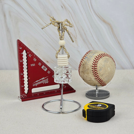 A red triangular ruler, a baseball on a stand, a black and yellow tape measure, and Jarhead Aromatics’ The Lumberjack Car Diffuser (Cedar, Oak & Mahogany) are arranged on a light surface against a marbled background.