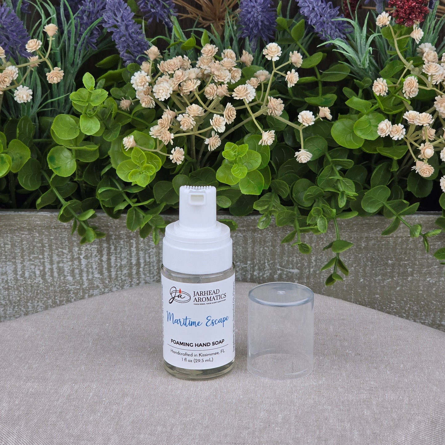 A bottle of Jarhead Aromatics Maritime Escape Foaming Hand Soap with the cap off sits on a beige surface in front of artificial green and purple floral arrangements.