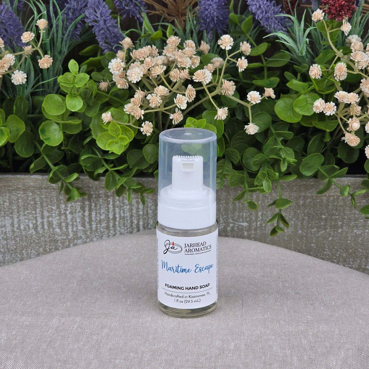 A bottle of Jarhead Aromatics Maritime Escape Foaming Hand Soap sits on a beige surface, backed by artificial green leaves and purple, white, and red flowers.