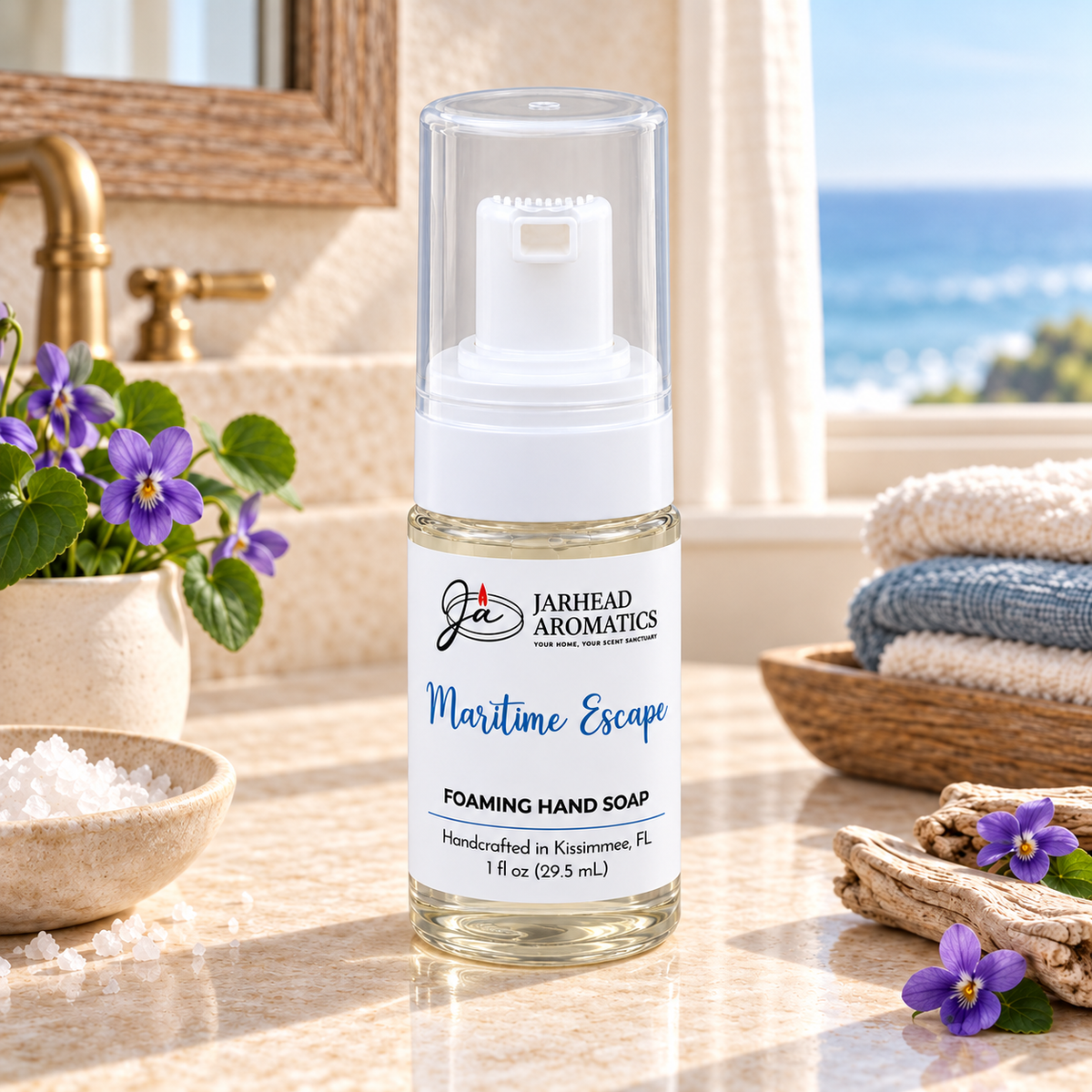 A clear bottle of Maritime Escape Handmade Foaming Hand Soap | Ozone, Sea Salt & Violet sits on a bathroom counter alongside purple flowers and sea salt, with a view of the ocean in the background.