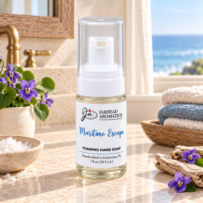 A clear bottle of Maritime Escape Handmade Foaming Hand Soap | Ozone, Sea Salt & Violet sits on a bathroom counter alongside purple flowers and sea salt, with a view of the ocean in the background.