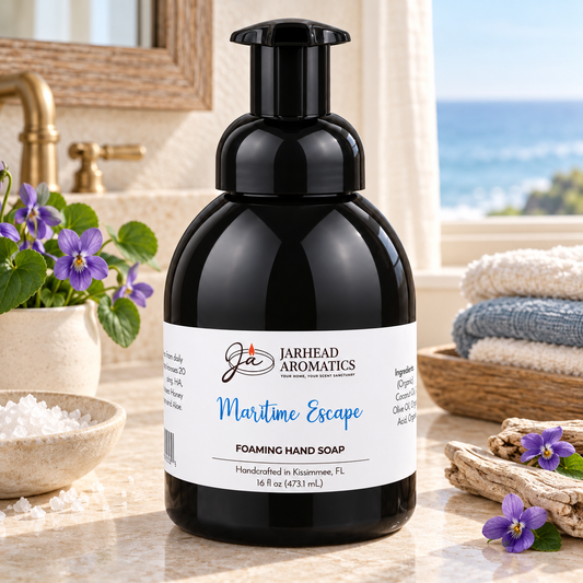 A black bottle of Maritime Escape Handmade Foaming Hand Soap with a white label, standing on a bathroom counter next to small purple flowers and folded towels.