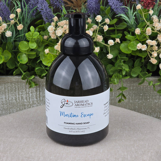 A bottle of Jarhead Aromatics Maritime Escape Foaming Hand Soap rests on a gray surface, backed by vibrant artificial flowers such as lavender and white blooms.