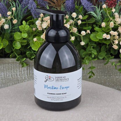 A bottle of Jarhead Aromatics Maritime Escape Foaming Hand Soap rests on a table, backed by vibrant artificial flowers and lush greenery.