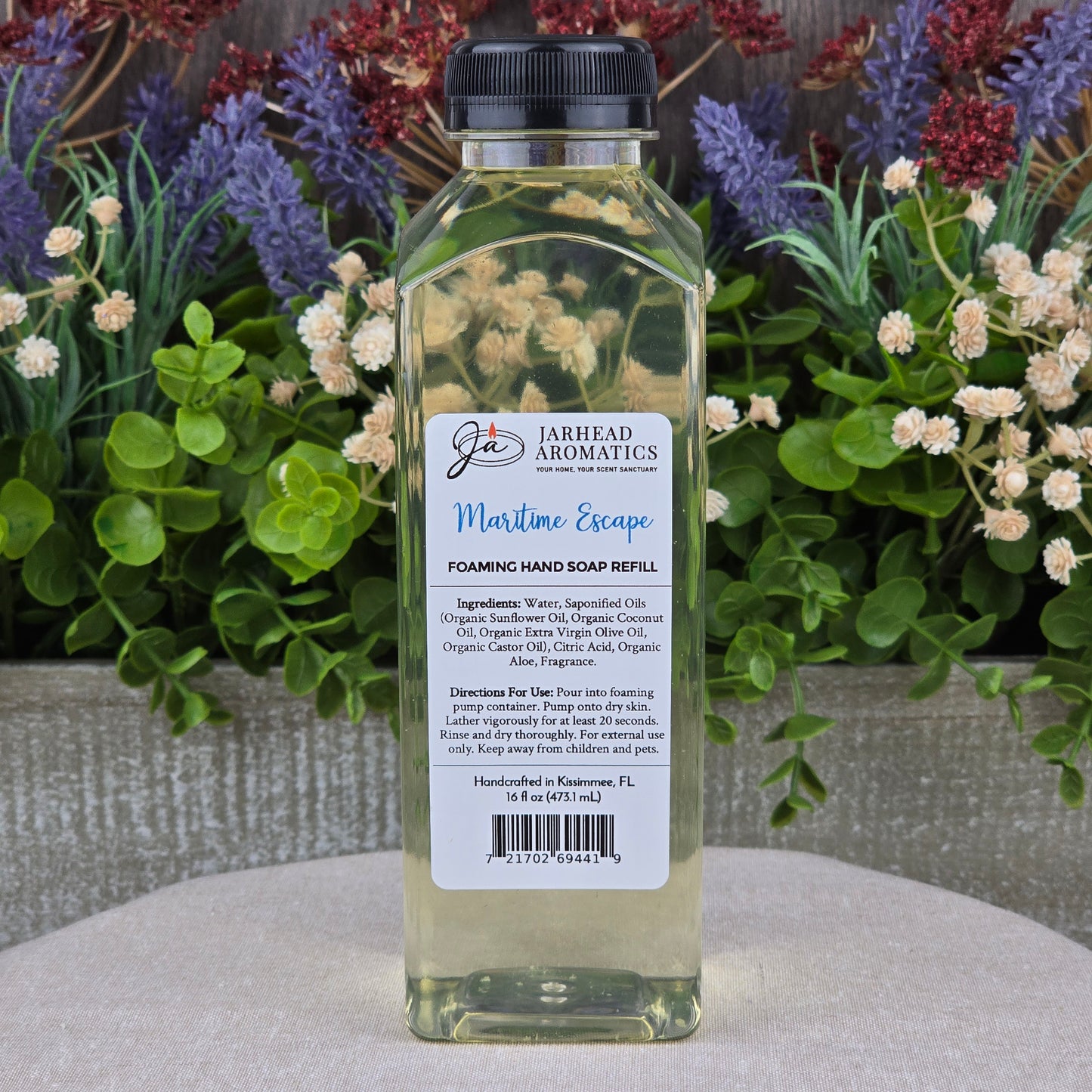 A clear plastic bottle of Jarhead Aromatics Maritime Escape Foaming Hand Soap sits on a round stone surface, surrounded by artificial flowers in purple, red, and green.