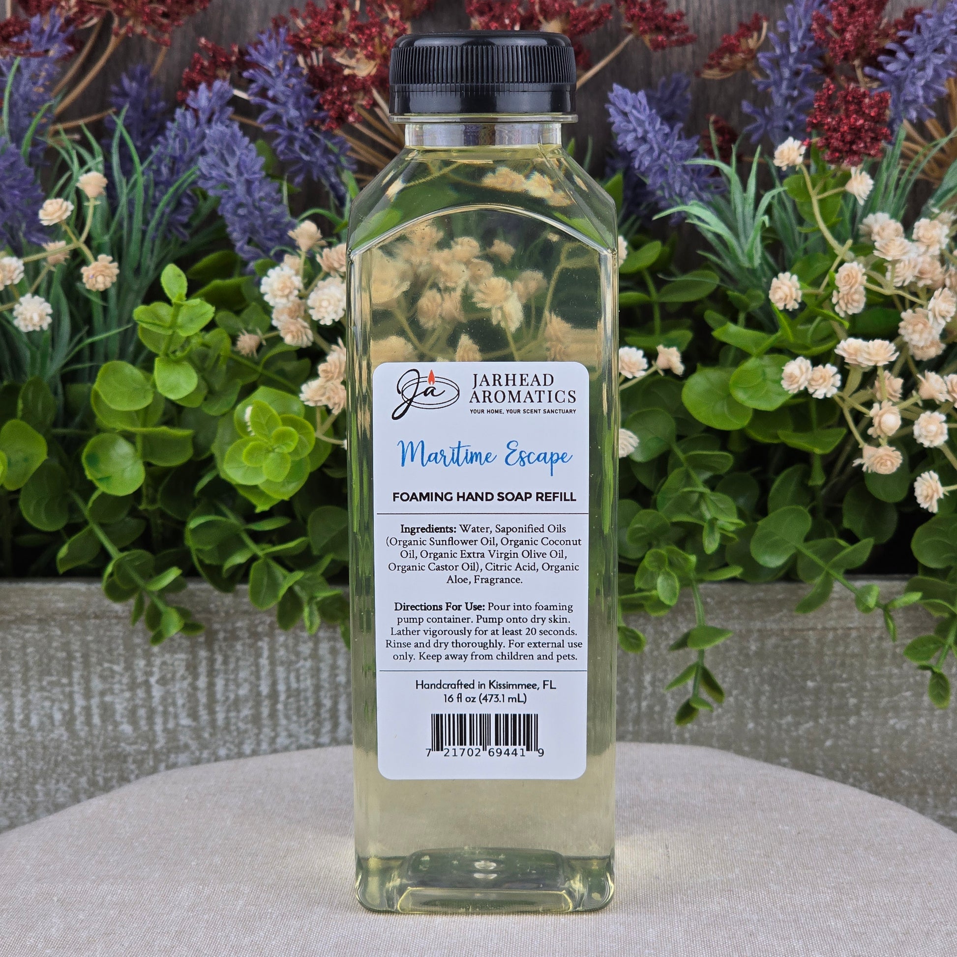 A clear plastic bottle of Jarhead Aromatics Maritime Escape Foaming Hand Soap sits on a round stone surface, surrounded by artificial flowers in purple, red, and green.