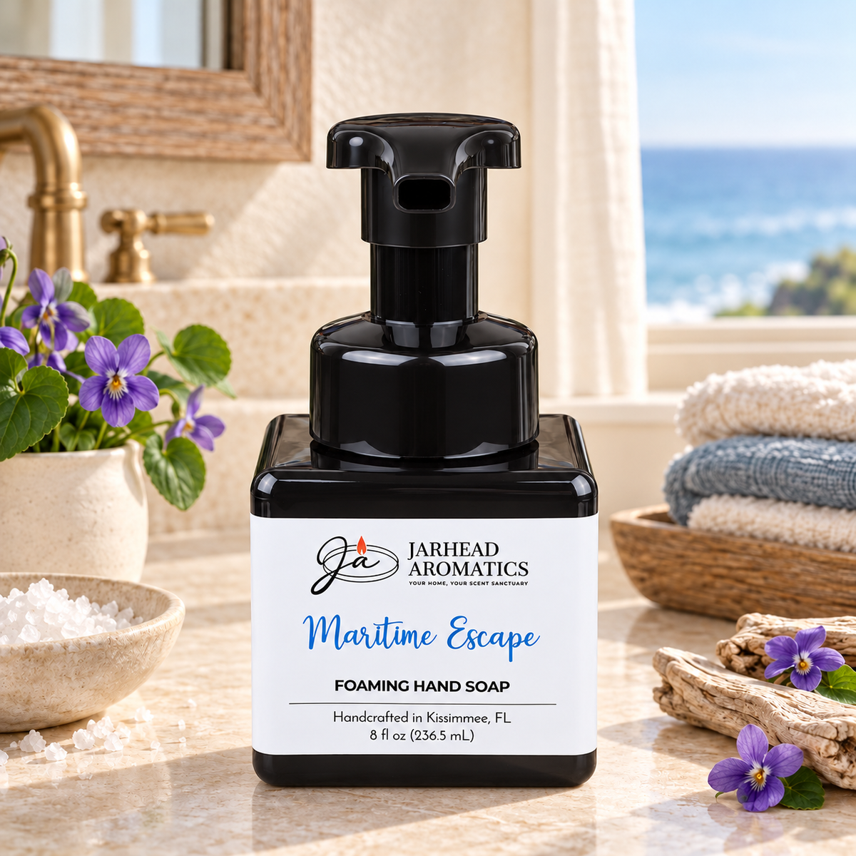 A square black bottle of Jarhead Aromatics Maritime Escape Handmade Foaming Hand Soap sits on a marble counter next to small purple flowers, a bowl of sea salt, and folded towels.