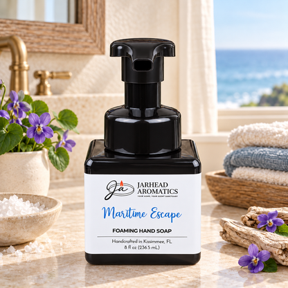 A square black bottle of Jarhead Aromatics Maritime Escape Handmade Foaming Hand Soap sits on a marble counter next to small purple flowers, a bowl of sea salt, and folded towels.