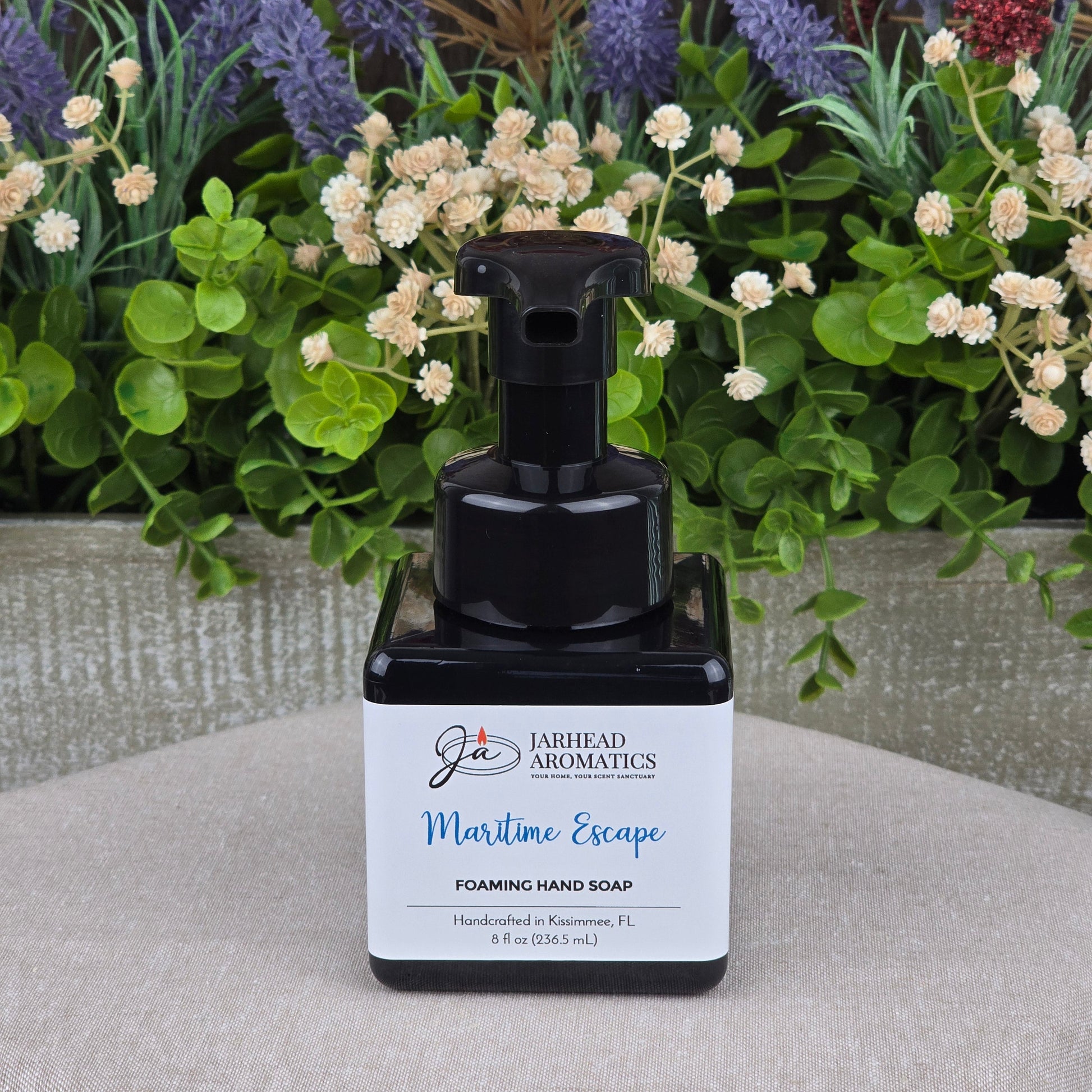 A black pump bottle of Jarhead Aromatics Maritime Escape Foaming Hand Soap rests on a beige surface, with vibrant artificial flowers and green leaves in the background.